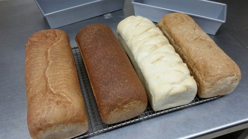 Four loaves of bread on a cooling rack, featuring different textures and colors, with baking pans in the background.