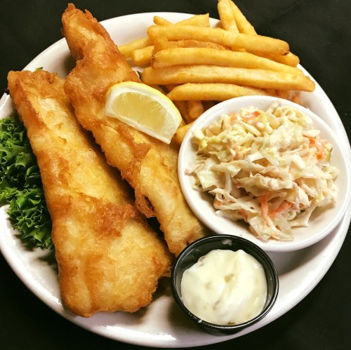 A plate of crispy fried fish fillets with lemon, fries, coleslaw, and a side of tartar sauce on a bed of lettuce.
