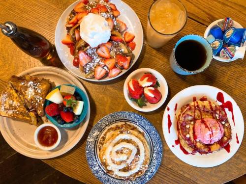 A table spread with various breakfast dishes: French toast, pancakes with strawberries, fruit, and drinks.