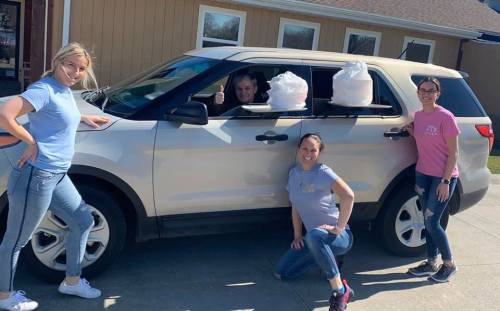 Four people pose by a car with bags in the windows, smiling and giving a thumbs up on a sunny day.