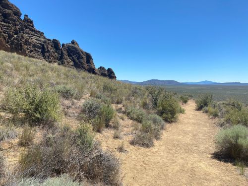 A dirt path winds through grassy terrain with rocky formations and distant mountains under a clear blue sky.