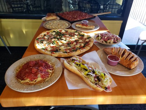 A table filled with various dishes: pizza, spaghetti, a sub sandwich, calzones, and a salad, all ready to eat.