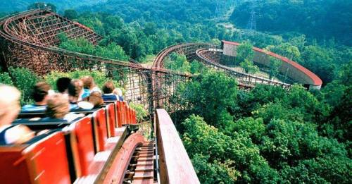 A wooden roller coaster winds through a lush green landscape, with riders enjoying the thrilling descent.