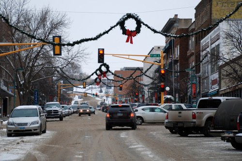 A snowy street scene decorated with holiday garlands, featuring cars and traffic lights in a small town.