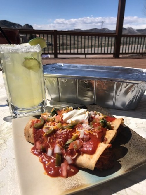 A plate of enchiladas topped with sauce, onions, and sour cream, alongside a margarita and a takeout container.