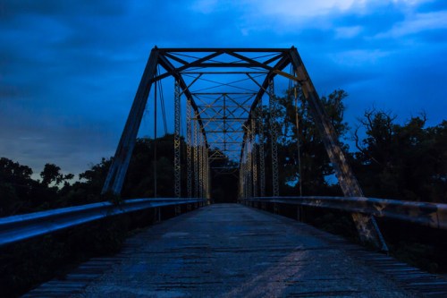 A dark, atmospheric view of a metal truss bridge at night, surrounded by trees under a cloudy sky.