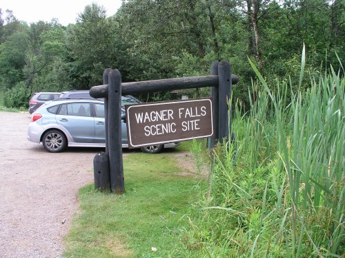 Sign for Wagner Falls Scenic Site, surrounded by greenery and parked cars in a natural setting.