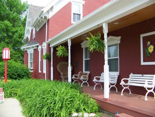 A charming red house with a porch, featuring white benches, hanging plants, and lush green landscaping.