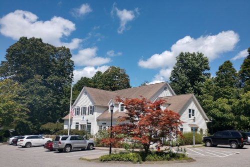 A white building with a sloped roof surrounded by trees and a parking lot under a blue sky with fluffy clouds.