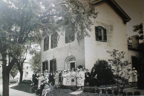 A large group of women in vintage clothing and hats gathered outside a historic building, surrounded by trees and greenery.