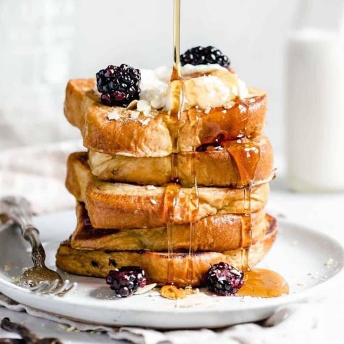 A stack of golden French toast topped with blackberries, syrup, and a dollop of cream on a white plate.