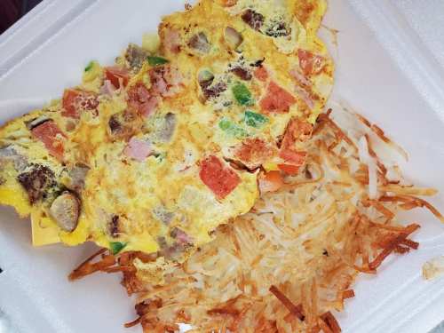 A colorful omelette with vegetables and ham served with crispy hash browns in a takeout container.