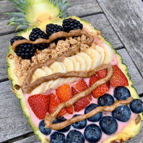 A pineapple half topped with granola, banana slices, strawberries, blueberries, blackberries, and drizzled with nut butter.