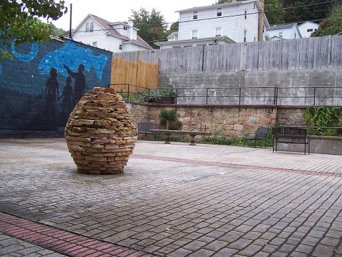A stone sculpture in a paved park area, with a mural and benches in the background.