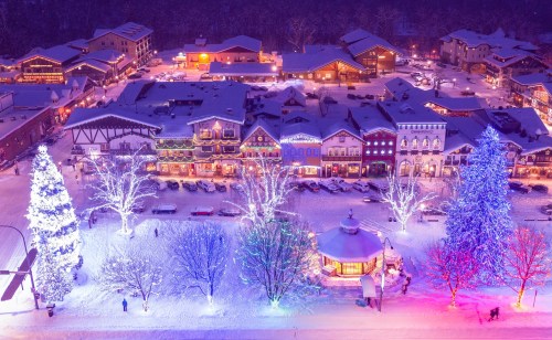 A snowy village scene at night, featuring colorful lights on trees and buildings, creating a festive atmosphere.