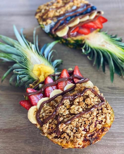 Two halved pineapples filled with granola, banana slices, strawberries, and drizzled with chocolate and berry sauce.