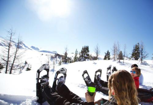 A group of people sitting in the snow, wearing snowshoes, enjoying a sunny winter day in a snowy landscape.