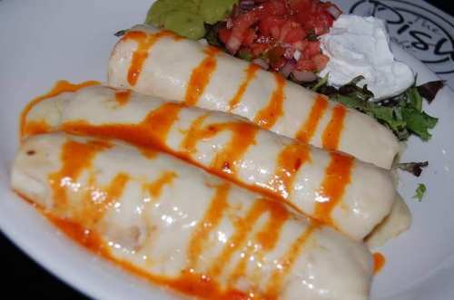 Three rolled enchiladas topped with cheese sauce and drizzled with red sauce, served with guacamole, salsa, and sour cream.