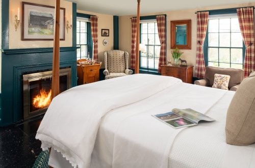 Cozy bedroom with a four-poster bed, fireplace, and large windows, featuring warm decor and a magazine on the bed.