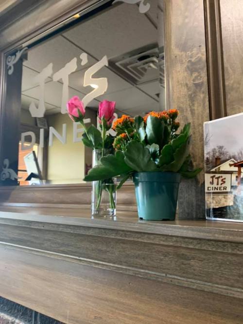 A wooden shelf with a mirror reflecting "JT's Diner," featuring pink roses and a potted plant.