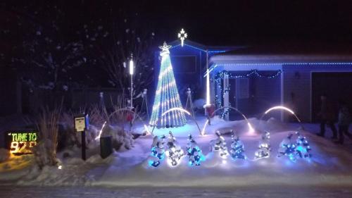 A snowy yard at night featuring a lit Christmas tree and colorful decorations, with a sign that says "Turn to 94.7."