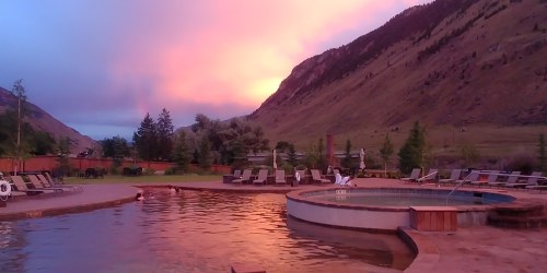 A serene pool area at sunset, surrounded by mountains and trees, with lounge chairs and a warm glow in the sky.