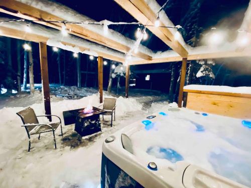 A cozy outdoor hot tub area with string lights, surrounded by snow and a fire pit, creating a warm winter atmosphere.
