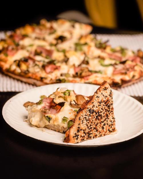 A slice of pizza with sesame seed crust on a plate, with a whole pizza in the background.