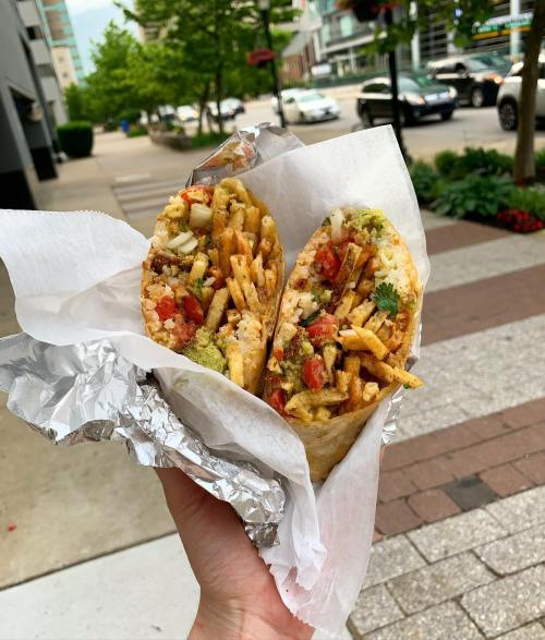 A hand holding two large burritos filled with meat, vegetables, and fries, wrapped in paper, on a city sidewalk.