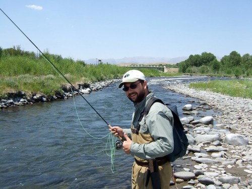 A man in fishing gear stands by a river, holding a fishing rod with a smile, surrounded by greenery and rocks.