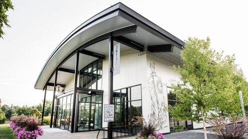 Modern building with large windows, curved roof, and greenery, featuring a mural and colorful flowers in the foreground.