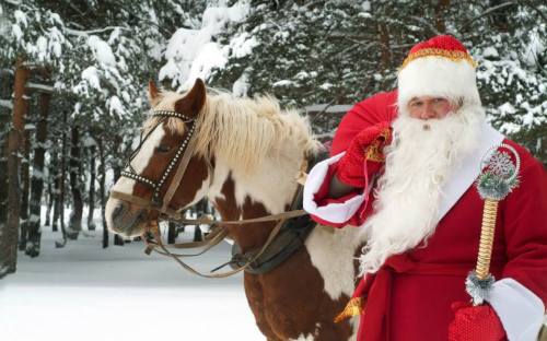 A Santa Claus figure in a red suit stands beside a horse in a snowy forest, holding a festive staff.