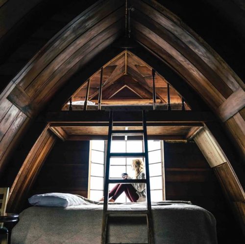 A cozy attic room with wooden beams, featuring a bed and a person sitting by the window, holding a cup.