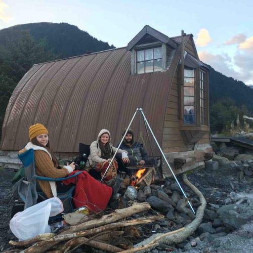 Three people sit by a campfire in front of a unique, curved cabin surrounded by mountains and trees.