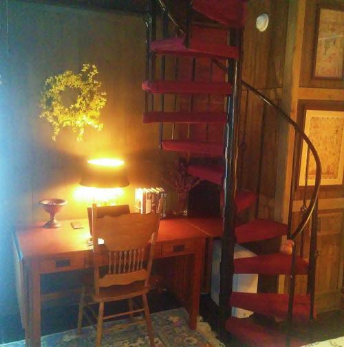 A cozy corner with a wooden desk, chair, and a spiral staircase, illuminated by a warm lamp and decorated with flowers.