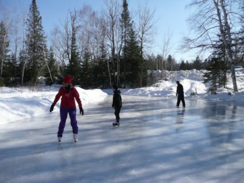 Three people ice skating on a frozen pond surrounded by snow-covered trees on a sunny day.