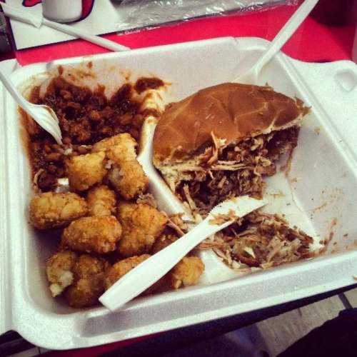 A takeout container with pulled pork sandwich, tater tots, and baked beans, served with plastic utensils.