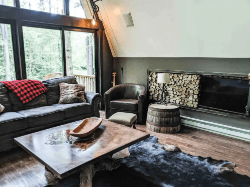 Cozy living room with a leather sofa, wooden coffee table, and a rustic fireplace, surrounded by large windows.