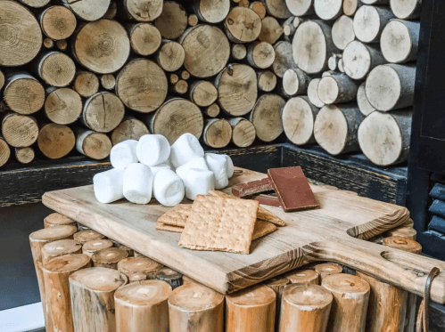 A wooden board with graham crackers, chocolate, and marshmallows, set against a backdrop of stacked logs.