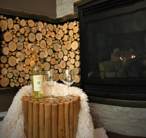 A cozy setting with a bottle of wine and glasses on a wooden table, next to a fireplace and a wall of stacked logs.