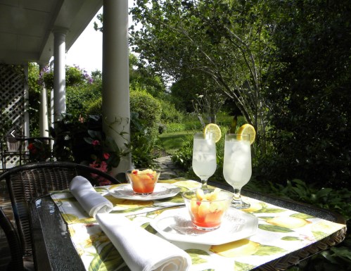 A sunny outdoor table set with two cocktails and a fruit dessert, surrounded by lush greenery and flowers.