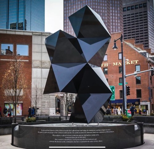 A large, abstract black sculpture stands in an urban area, surrounded by buildings and people.