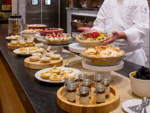 A chef presents a colorful fruit tart at a dessert display featuring various pies and pastries on a countertop.