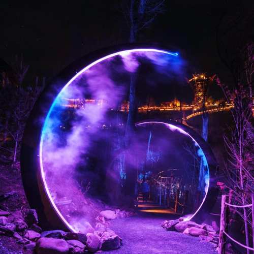 A glowing circular archway with purple lights in a dark forest, leading to a distant illuminated structure.
