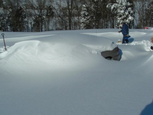 A car is completely buried under a thick layer of snow, with only its outline visible in a winter landscape.