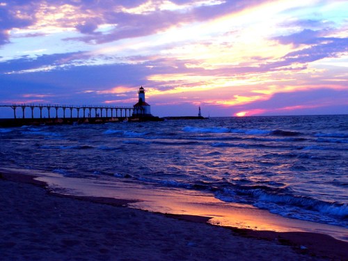 A serene beach at sunset, featuring a lighthouse and a pier silhouetted against colorful skies and gentle waves.