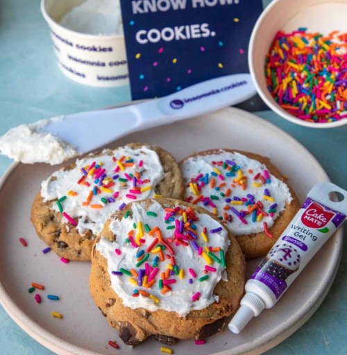 Three frosted cookies topped with colorful sprinkles, a tube of writing gel, and a bowl of sprinkles in the background.
