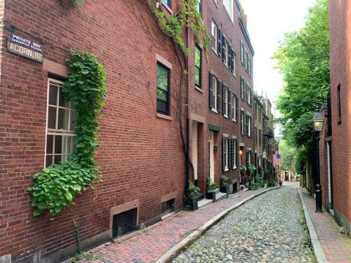 A narrow, cobblestone street lined with brick buildings and greenery, featuring a sign for Acorn Street.