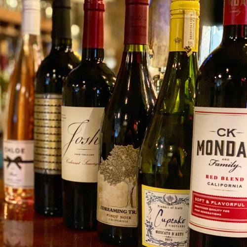 A selection of wine bottles lined up on a bar, featuring various labels and colors, including reds and a white.