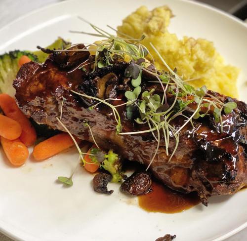 A plated dish featuring a glazed meatloaf, garnished with microgreens, served with carrots, broccoli, and mashed potatoes.